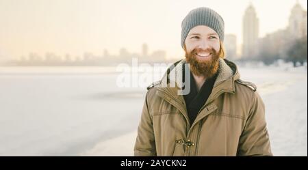 Close-up Caucasian young male red hair and beard in a hat and a park coat posing winter model against a background of a lake to Stock Photo