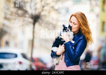 A young redhaired Caucasian woman with freckles holds and kisses, embracing black and white shaggy dog of Chihuahua breed. girl Stock Photo