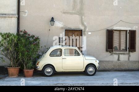 Small vintage italian car. Beige color old car in front of old house facade and flowers.. Stock Photo