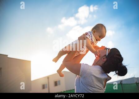 An emotional picture of 1 year old baby and her mother holding her up in the air against blue sky and bright sun Stock Photo