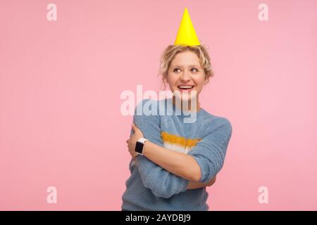 Portrait of glad carefree adult woman in warm sweater and funny cone hat on head smiling and embracing herself, looking excited about entertainment, b Stock Photo