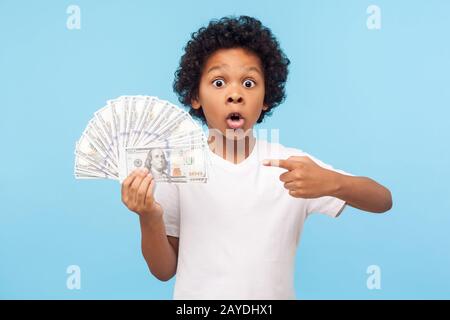 Wow, I'm rich! Amazed lucky wealthy little boy with curly hair pointing to fan of dollar banknotes, shocked by lots of cash and looking at camera with Stock Photo