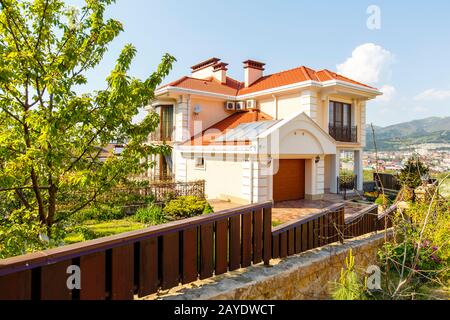 The facade of a beautiful white two-story cottage with a red roof. The cottage is located behind a wooden fence made of dark lacquered wood between st Stock Photo