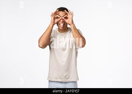 Guy popping eyes and looking amused through finger glasses. Happy carefree handsome man in t-shirt, showing goggles stick tongue Stock Photo