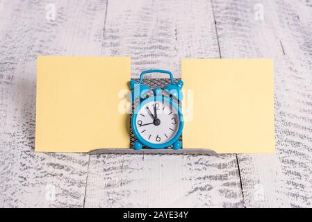 Mini blue colour alarm clock to show time placed in between two yellow notepapers. Classic little watch standing above a buffer