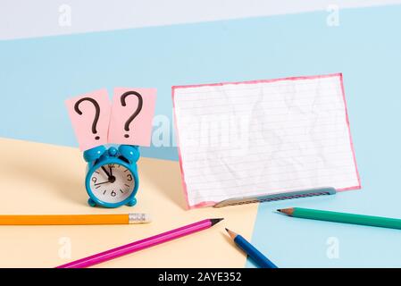 Mini small size alarm clock beside stationary placed tilted. Notepaper on the edge of empty plain multi colours table backdrop. Stock Photo