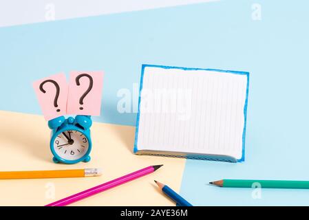 Mini small size alarm clock beside stationary placed tilted. Notepaper on the edge of empty plain multi colours table backdrop. Stock Photo