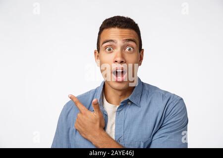 Enthusiastic, surprised hispanic man in blue shirt over t-shirt, pointing upper left corner, gasping and drop jaw amazed, poppin Stock Photo