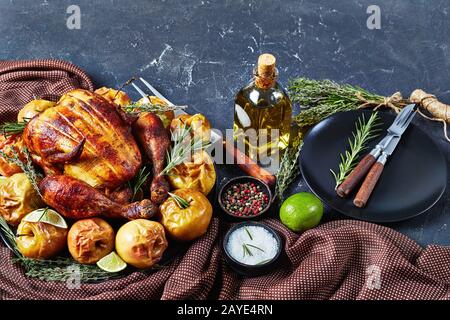 close-up of a whole rotisserie chicken served on a black platter with baked apples and aromatic herbs on a concrete table with fork and spices, horizo Stock Photo