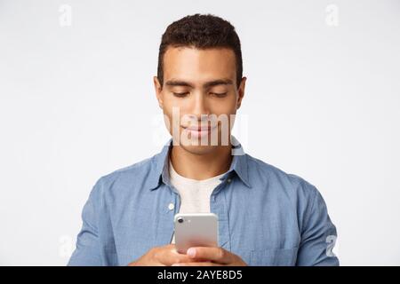 Handsome stylish modern hispanic man in casual outfit, holding smartphone, smiling as typing message to girlfriend, scrolling so Stock Photo