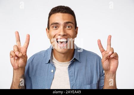 Surprised, handsome masculine young hispanic guy in shirt over t-shirt. showing peace or quotation gesture, smiling astonished, Stock Photo