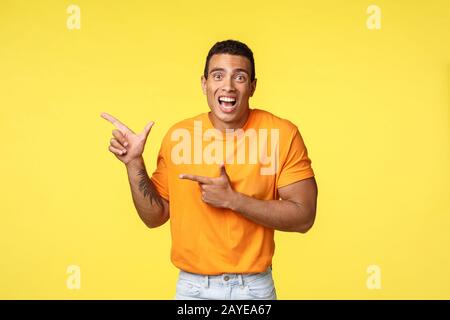 Cheerful excited man with tattooed arm in orange t-shirt, smiling astonished telling something amazing happened pointing left to Stock Photo