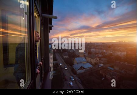 Adult man stands on the balcony and looks into the distance enjoying the sunset. Beautiful city landscape of a fiery sky at sunset. Roofs of houses and passing cars in the bustle of the city Stock Photo