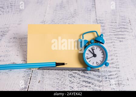 Mini blue colour alarm clock to show time placed tilted in front of one yellow notepaper. Classic little watch standing above a Stock Photo