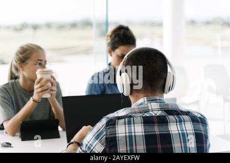 Back of a man with white helmets sitting next to two girls at the same table meeting and working together in a coworking with laptops and coffee Stock Photo
