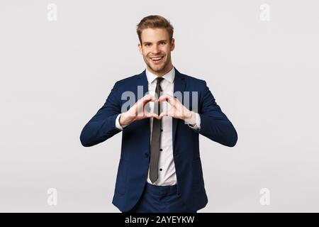 Joyful bearded young blond man in blue classic suit, showing heart sign and smiling, come from office home, make surprise dinner Stock Photo