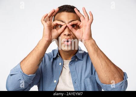 Surprised and amused funny young handsome man making superhero mask or glasses with fingers, folding lips from amazement, impres Stock Photo