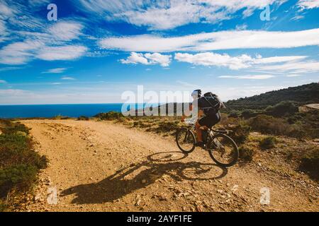 a young guy riding a mountain bike on a bicycle route in Spain on road against the background of the Mediterranean Sea. Dressed Stock Photo