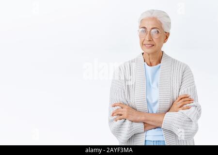 Thoughtful elderly woman in glasses, warm comfortable plaid cadrigan, cross arms chest, smiling and looking left interested, entertained, watching tv Stock Photo