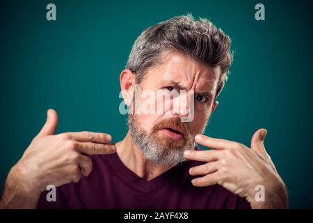 A portrait of bearded surprised man in red t-shirt pointing fingers at himself. People and emotions concept Stock Photo