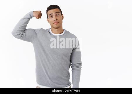 Confident and cheeky young hispanic athletic man, bragging with body strength, flex biceps and smiling, showing muscles, smirk boastful, standing Stock Photo