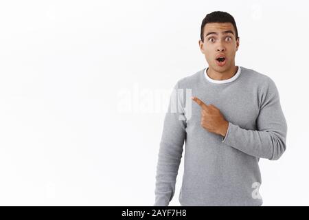 Surprised and impressed, startled guy in grey sweater, react to interesting news, pointing finger upper left corner, telling about cool event going Stock Photo