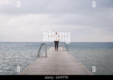 Concept of freedom relaxation. woman relaxing on pier. Woman hiker backpacker with backpack on sea wooden pier goes forward. Emo Stock Photo