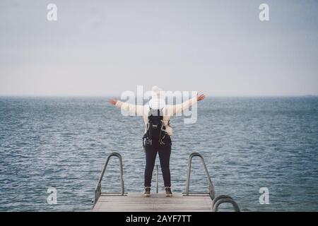 Concept of freedom relaxation. woman relaxing on pier. Backpacker hiker woman with backpack on sea wooden pier stands arms sidew Stock Photo