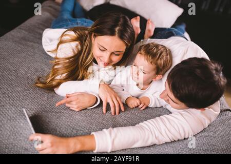 A young family lies on the bed and looks at the mobile phone. Mom, dad and son are watching a video on a smartphone in the bedro Stock Photo