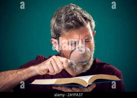 A portrait of bearded man in red t-shirt holding opened book and magnifying glass. People, education and searching concept Stock Photo