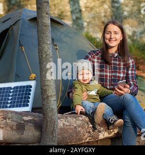 Mother and son are sitting on a log near the solar panel and tent in the forest on a sunny day. Cute baby boy looking at the camera with a smile, a woman holding a mobile phone in her hand Stock Photo