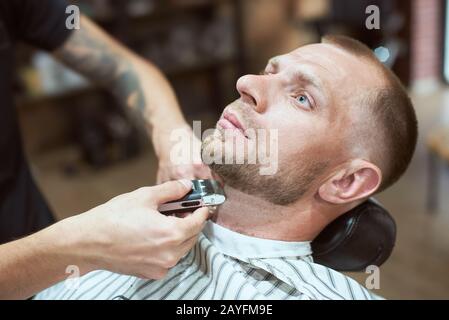 Getting perfect shape. Close-up side view of young bearded man getting beard haircut by a tattooed hairdresser at barber shop Stock Photo