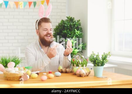 Happy easter.A funny fat man decorates eggs while sitting at a table with Easter decor in the background Stock Photo