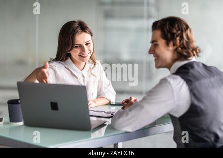 Beautiful business woman and handsome businessman in formal suits are using a laptop, discussing documents and smiling while working in office Stock Photo
