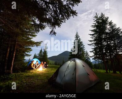 Group of tourists basking by campfire near pine forest with spectacular mountain landscape in evening. Green trees and grassy hills under sky after sunset, on which stars appear between light clouds Stock Photo