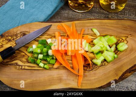 Close up of isolated sliced fresh spring onions (scallions), carrots, cucumbers on wooden olive tree cutting board and japanese kitchen knife. Blue na Stock Photo