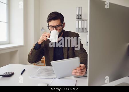 Businessman worker drinking a coffee mug while sitting at a table with a computer working in the office. Stock Photo