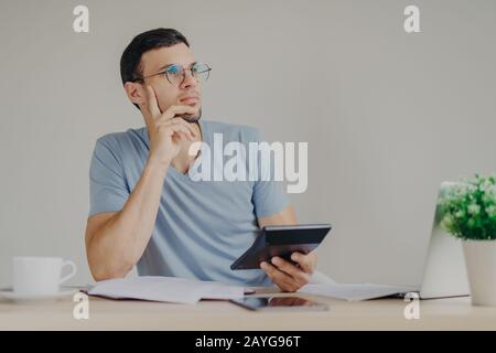 Serious pensive male wears round glasses calculates expanses uses generic laptop computer and calculator, surrounded with mug of coffee and documentat Stock Photo