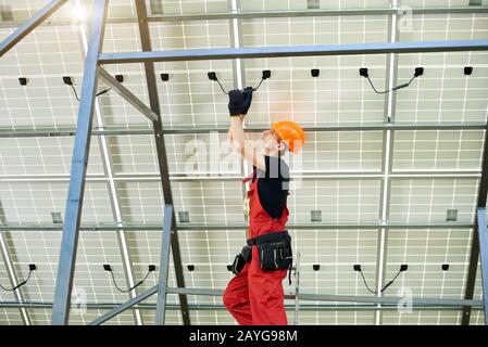 Male expert inspecting quality of a solar panels. Worker in orange uniform and helmet with equipment. Ecology power conservation concept. Stock Photo