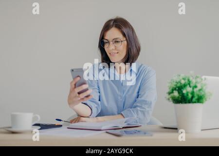 Successful brunette female freelancer recieves good news on smart phone while works on laptop computer, reads post on website, makes notes in diary, d Stock Photo
