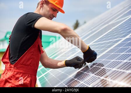 Electrician installing solar panels to save energy. A man is wearing an orange uniform, hard hat, black t-shirt and gloves against the blue sky. Alternative energy sustainable concept Stock Photo