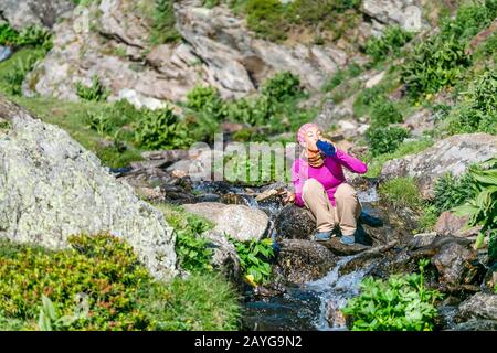 carefree woman hiker taking water from forest strean and drink it. Stock Photo