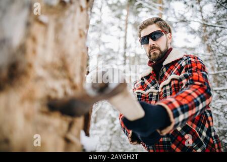 Handsome stylish man in checkered winter jacket chopping a tree with an axe Stock Photo
