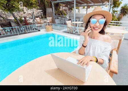 Happy asian woman wirking with mobile phone and laptop pc and relaxing near swimming pool. Distant work concept Stock Photo
