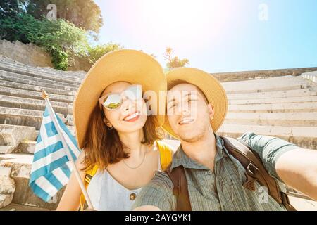 European man and Asian girl in love a couple of tourists take a selfie in an ancient Greek Acropolis with a flag. Travel and adventure in Greece Stock Photo