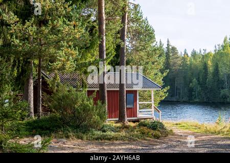 Red wooden finnish traditional cabins cottages in green pine forest near river. Rural architecture of northern Europe. Wooden houses in camping on sun Stock Photo