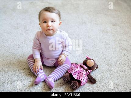 Thoughtful and serious nice baby girl sitting on the floor with toy bear at home Stock Photo