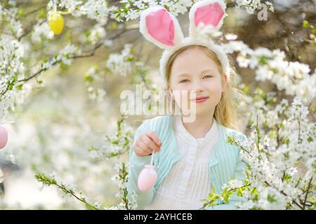 Adorable little girl wearing bunny ears in blooming cherry garden on beautiful spring day. Kid hanging Easter eggs on blossoming cherry branches. Chil Stock Photo