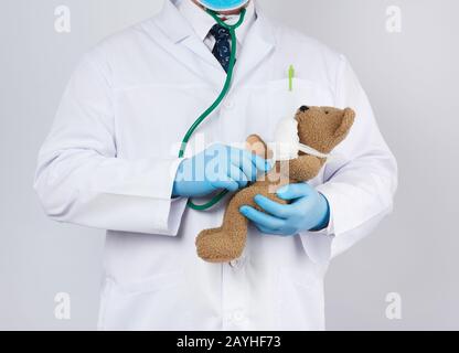 pediatrician in a white coat, blue latex gloves holds a brown teddy bear with a bandaged paw, doctor examines the toy with a stethoscope, white backgr Stock Photo