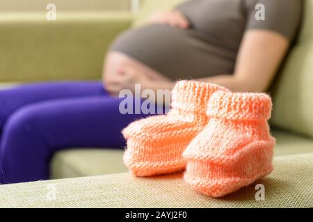 A beautiful woolen baby shoes on the couch next to a pregnant woman Stock Photo